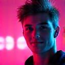 Headshot of an energetic queer DJ and event promoter smiling confidently against a blurred club background.